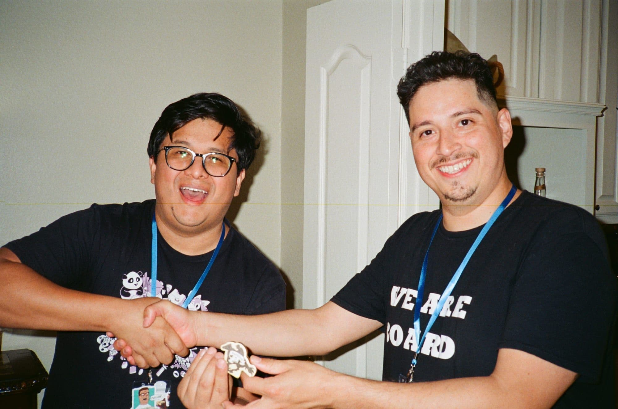 Two listeners in WeAreBoard shirts shaking hands over a meeple trophy